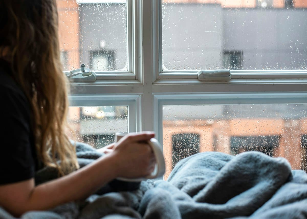 Persona con una taza en las manos mirando por una ventana en un día lluvioso
